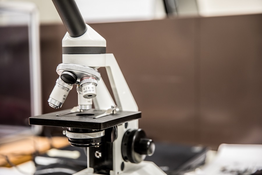 Man sieht eine Nahaufnahme von einem optischen Mikroskop. Es befindet sich kein Objektträger auf dem Objekttisch. Das Mikroskop ist im Fokus, die Objekte im Hintergrund des Tisches, auf dem das Mikroskop steh, sind verschwommen. 