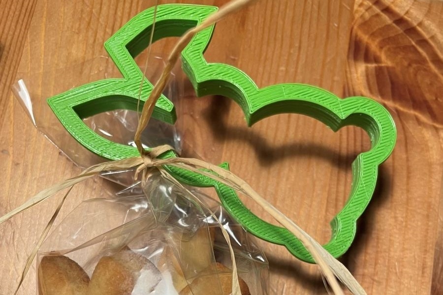 Man sieht in Cellophan verpackte Guetzli. Wo das Cellophan mit einer Masche verschnürt wurde, ist ein 3D-gedrucktes, grünes Förmchen in Form eines Osterhasen, dessen eines Ohr leicht geknickt ist, befestigt. 