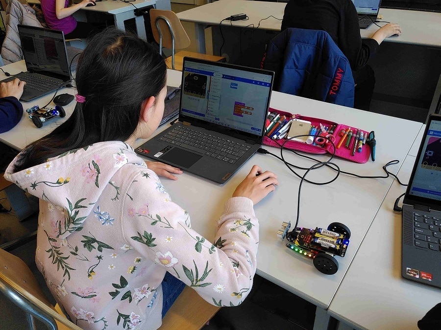 Auf dem Bild sieht man ein Mädchen vor einem Notebook sitzen. Auf dem Notebook ist eine grafische Programmieroberfläche zu sehen, mit welcher man den ebenfalls an den Computer angeschlossenen Roboter und dessen Sensoren (BitBot, Fahrzeug) programmieren kann. 