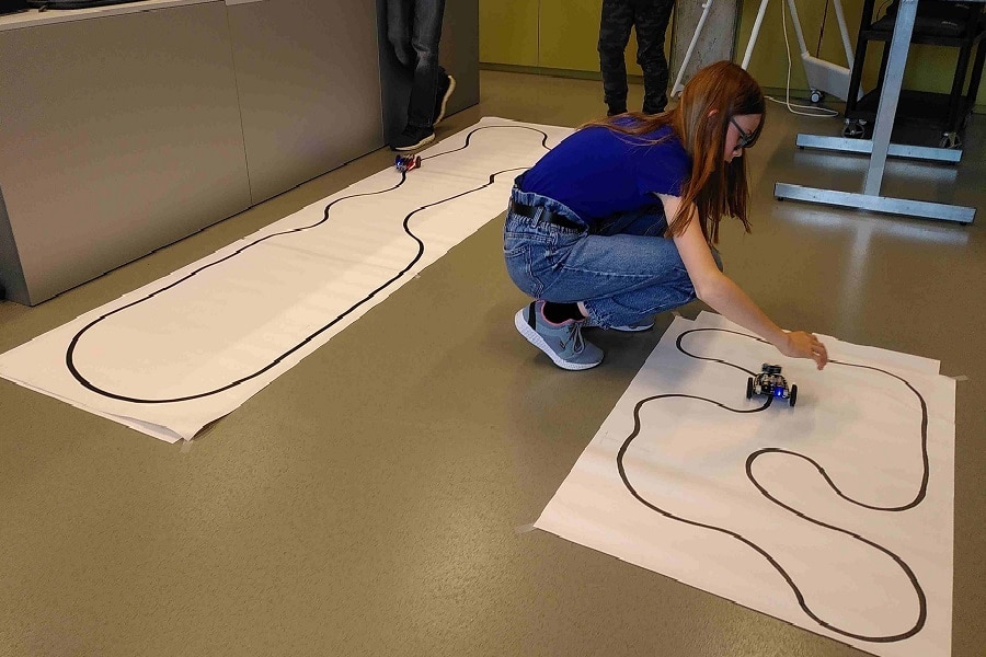 Auf dem Bild sieht man ein Mädchen, welches zwischen zwei auf grosse Bahnen Papier gemalten Autorennstrecken hockt. Sie setzt gerade ihren BotBit, das selbst programmierte Roboterfahrzeug, auf die eine, sehr kurvenreiche Strecke.