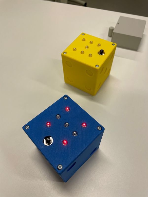 Man sieht einen blauen und einen gelben elektronischen Spielewürfel. Beim blauen leuchten vier Leuchtdioden und zeigen eine Vier an, der gelbe Würfel ist ausgeschaltet. 