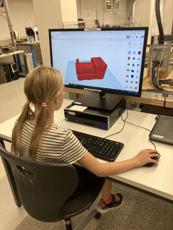 Auf dem Bild ist ein Mädchen am Computer zu sehen, welches eine rote Lokomotive in einem 3D-CAD-Programm konstruiert hat.