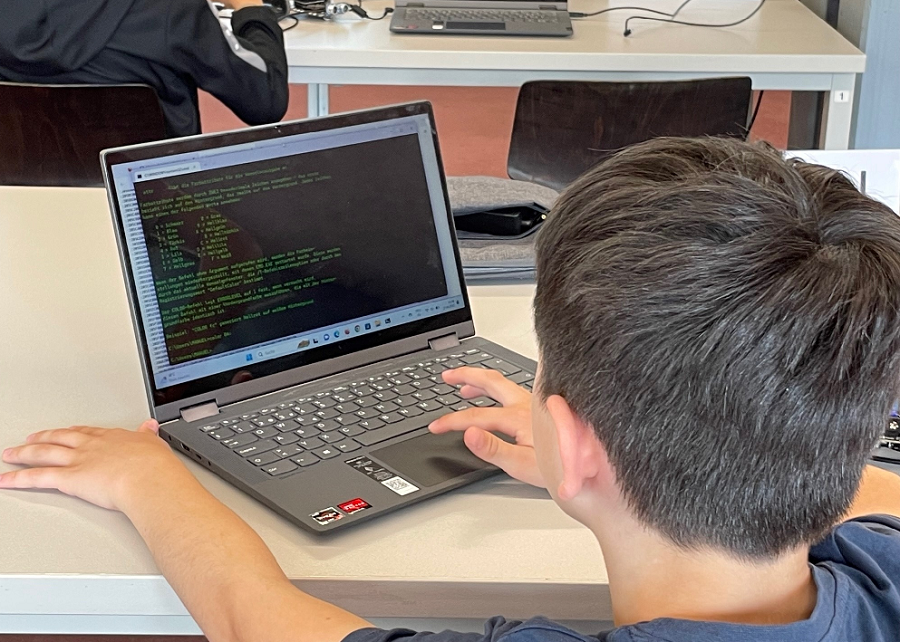 Auf dem Bild ist ein Junge von hinten zu sehen, der auf den Bildschirm eines Notebooks schaut, wo grüner Code auf schwarzem Hintergrund zu sehen ist. 