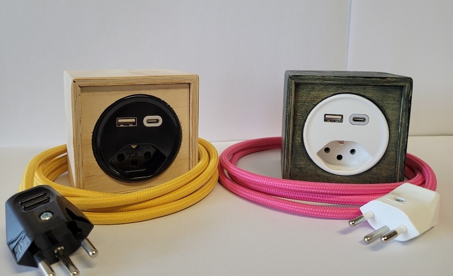 Man sieht zwei Power-Cubes. Einen hellen, mit einer schwarzen Steckdose sowie USB- und USB-C-Anschluss mit gelbem Kabel und schwarzem Stecker sowie einen zweiten, dunklen mit weisser Steckdose sowie USB- und USB-C-Anschluss mit pinkem Kabel u nd weissem Stecker. 